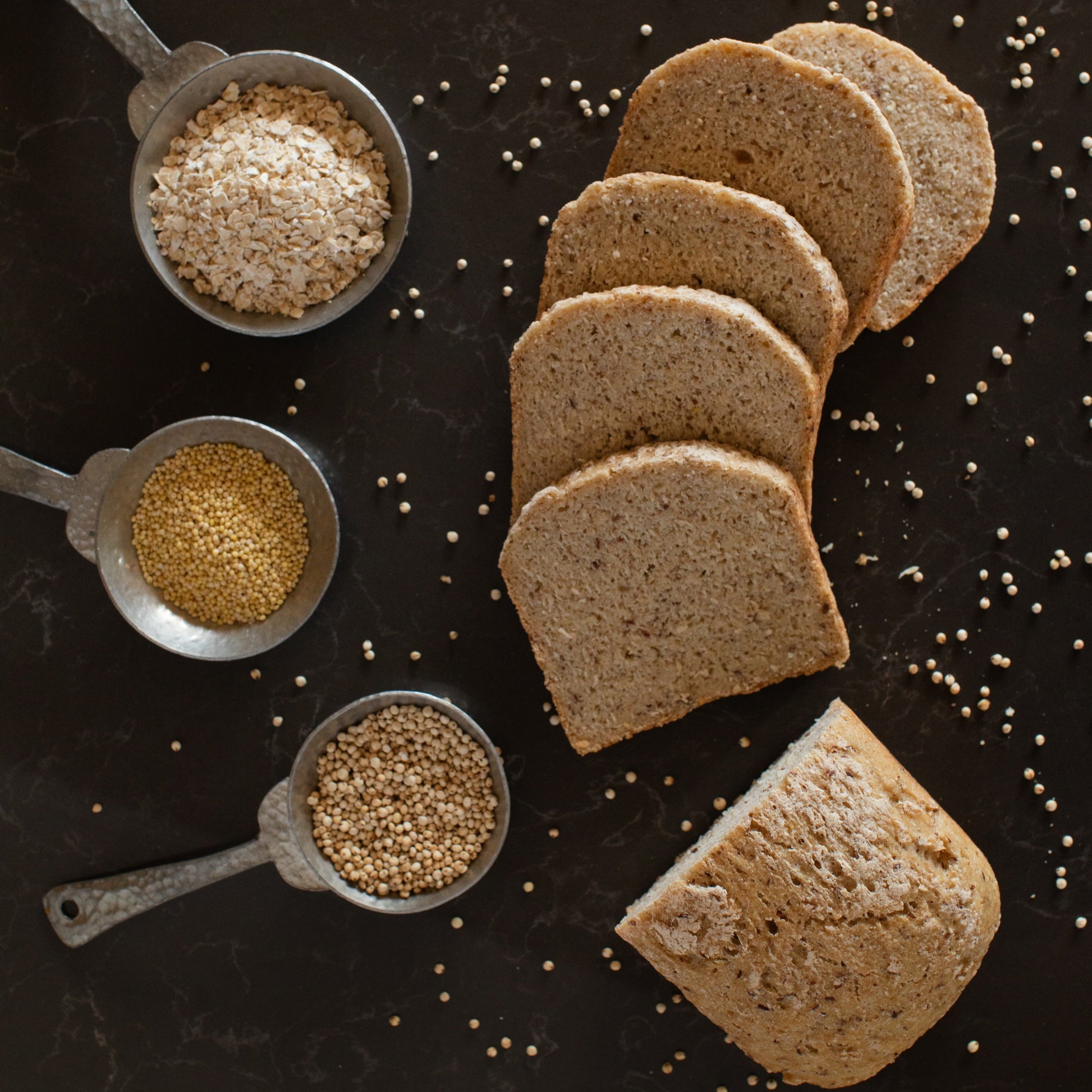 Sliced organic gluten-free sourdough bread next to measuring cups filled with grains and seeds, all arranged on a dark surface.